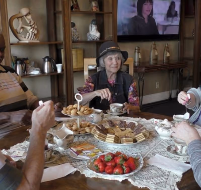 Seniors seated around a lace-covered table with tea cups, pastries, and a plate of strawberries in a cozy common room.