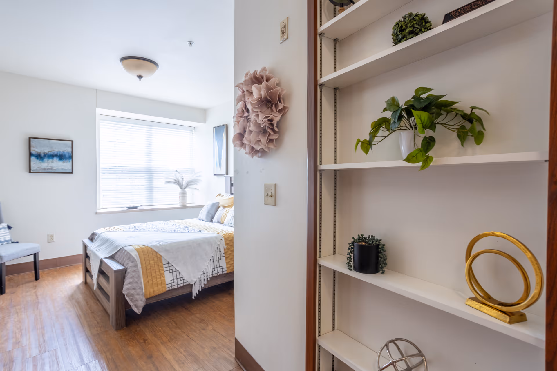 Bright furnished bedroom with a bed by a large window and built-in shelving with plants and decorative items in the foreground.