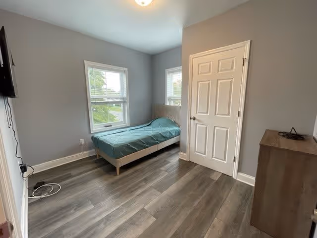 A small, simple bedroom with gray walls and wood flooring. The room contains a single bed with a green cover positioned near two windows, a closed white door, a wooden dresser, and a wall-mounted television.