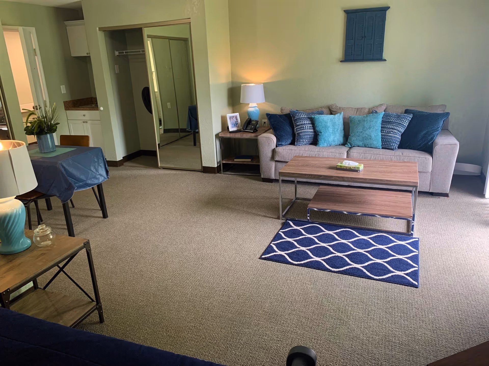 A cozy living room area in a retirement community featuring a beige sofa with multiple blue and teal cushions, a wooden coffee table with a tissue box on top, a small blue and white patterned rug, and a side table with a lamp and framed photo. There is a small dining table with chairs covered with a blue tablecloth, a closet with mirrored sliding doors, and a small kitchenette area visible in the background.