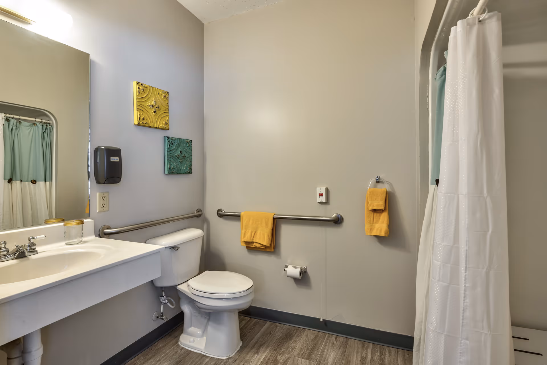 A bathroom with a white toilet, a white sink with a mirror above it, and a shower with a white curtain. The walls are light gray, and there are two decorative square wall hangings in yellow and teal. Yellow towels hang on metal grab bars and a towel ring. The floor has wood-like vinyl flooring.