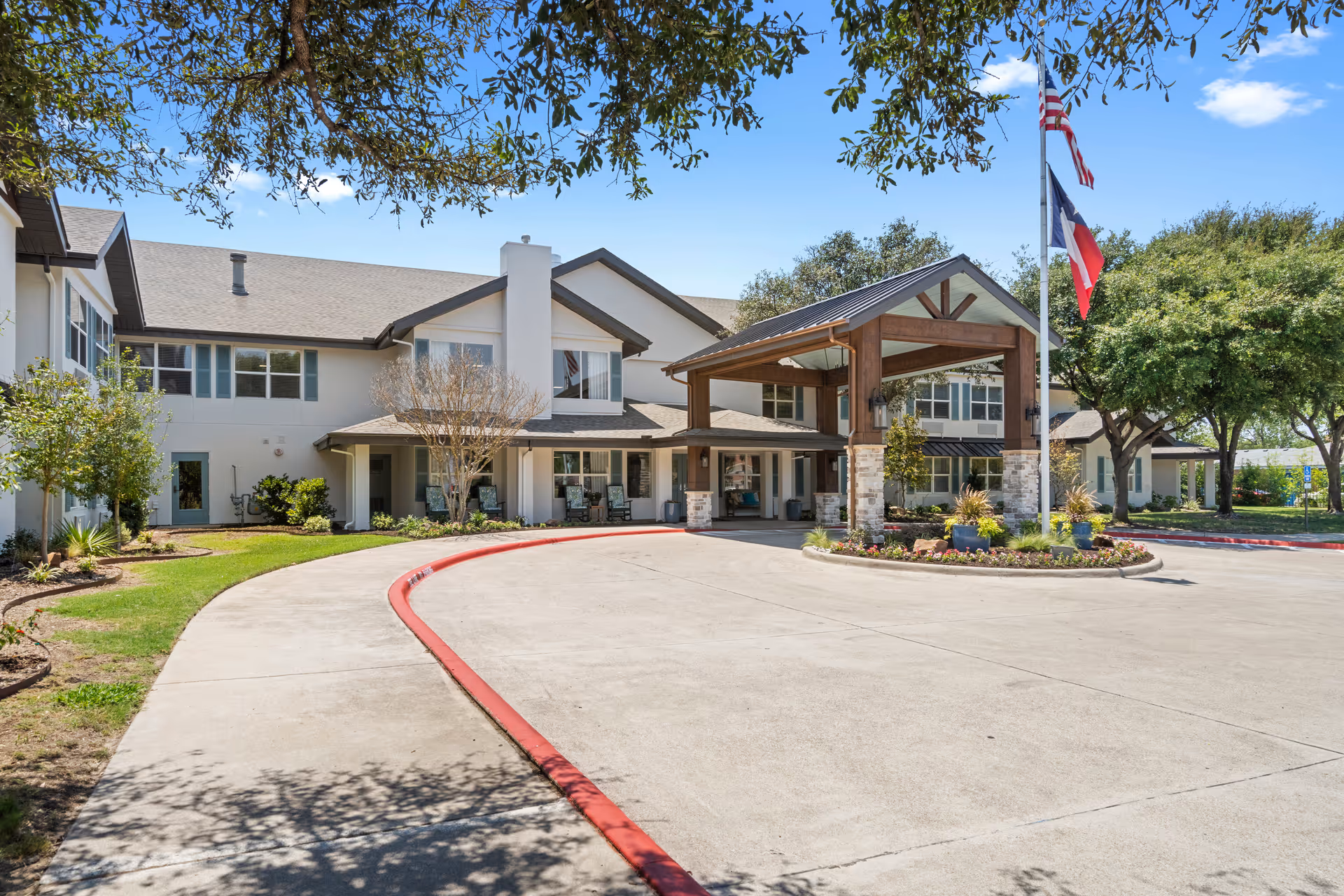 Front entrance of a senior living facility with a covered porte-cochere, flagpoles, landscaping, and a circular driveway.