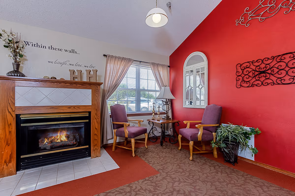 A cozy sitting area with a lit fireplace on the left, two purple upholstered wooden armchairs facing each other with a small wooden table and lamp between them, a large window with beige curtains behind the chairs, a red accent wall with decorative metal wall art and a white framed mirror, and a vase with flowers on the fireplace mantel. The wall above the fireplace has a quote that reads 'Within these walls... we laugh... we love... we live.'