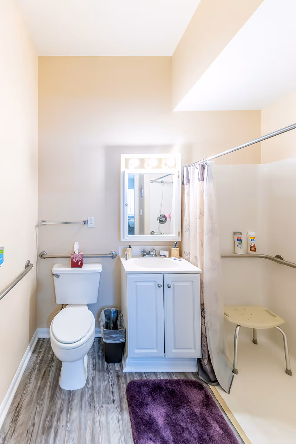 A clean and well-lit bathroom with beige walls and wood-look flooring. The bathroom features a white toilet with a tissue box on top, a white vanity with a sink and a mirror above it with three lights. There is a purple bath mat on the floor. To the right is a walk-in shower with a floral shower curtain, a shower chair, and grab bars installed on the walls for safety. A small trash can is placed between the toilet and the vanity.