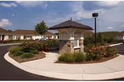 A landscaped roundabout with a stone and wood gazebo-like structure in the center, surrounded by flowers and shrubs. Single-story buildings with brown roofs are visible in the background under a partly cloudy sky.