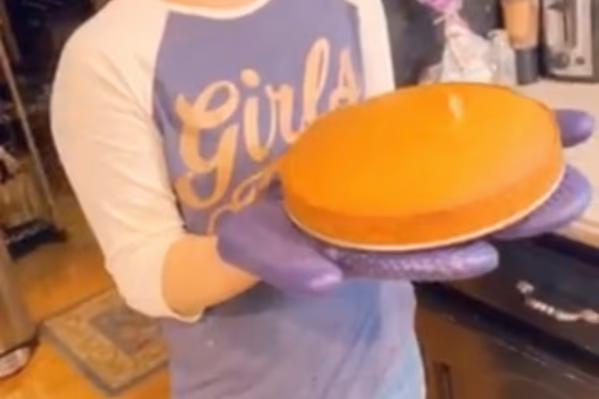 Person wearing purple gloves holding a round baked cheesecake in a kitchen setting.