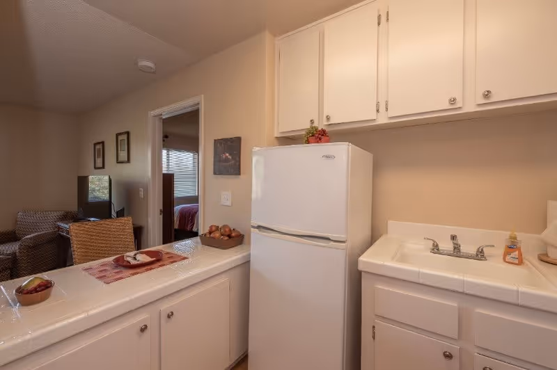 Interior view of a small kitchen area in a senior living facility with white cabinets, a white refrigerator, a sink with a faucet, and a countertop with a bowl of fruit and a decorative placemat. In the background, there is a glimpse of a living room with a chair and a television, and a bedroom with a bed and window blinds.