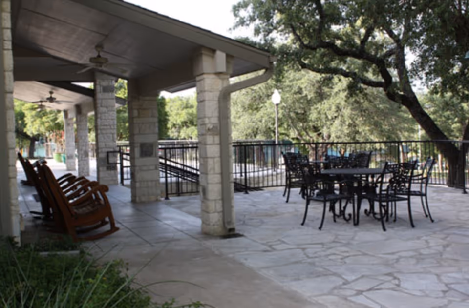 Outdoor covered patio area with stone pillars, ceiling fans, wooden rocking chairs on the left side, and black metal tables and chairs on the right side surrounded by trees and greenery.