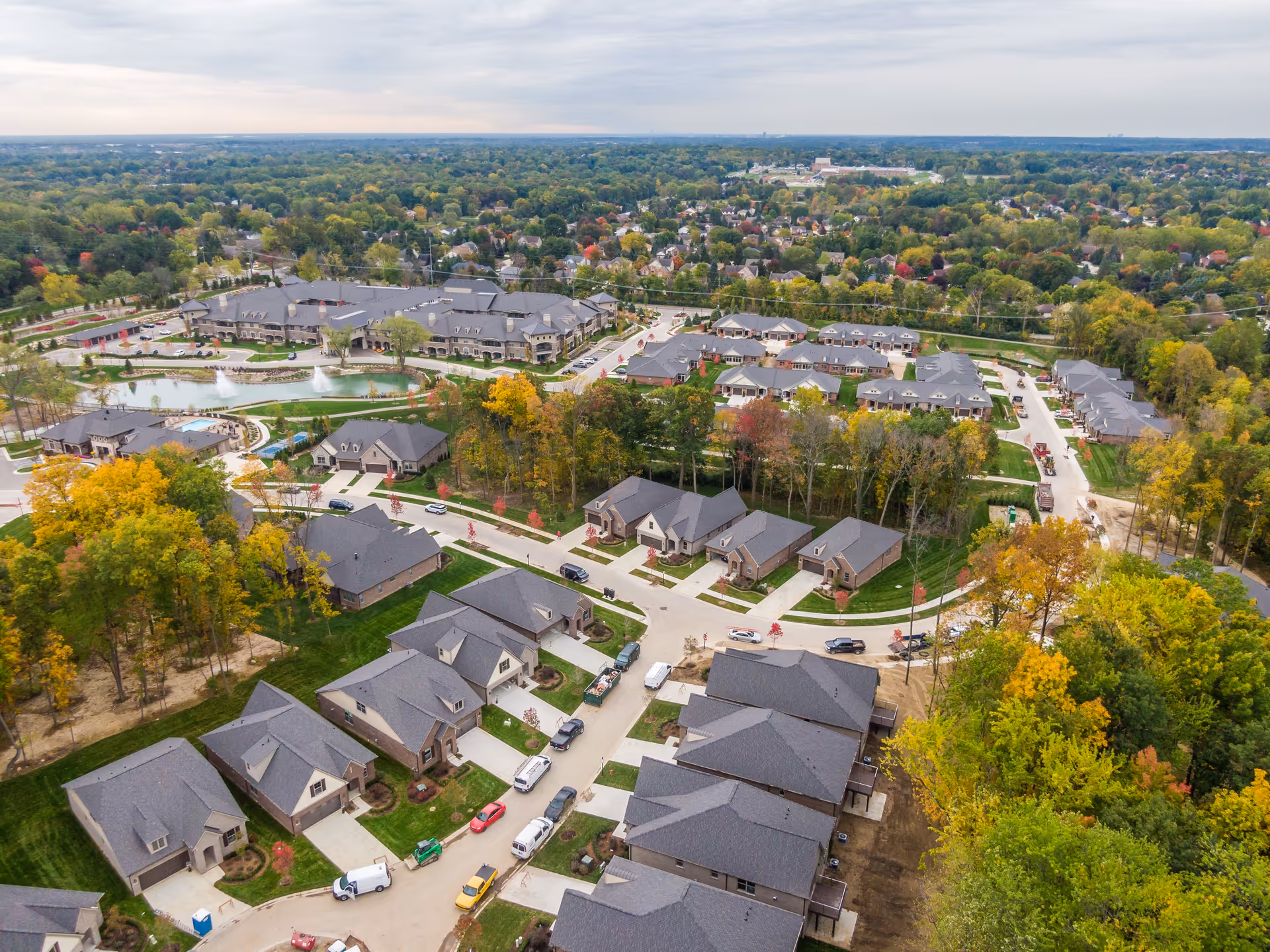 Aerial view of the Villages at Flourish Collection senior living community showing multiple residential buildings with gray roofs, surrounded by trees with autumn foliage. There is a pond with a fountain near the center, paved roads, parked vehicles, and a large building in the background. The landscape extends to a distant horizon under a cloudy sky.