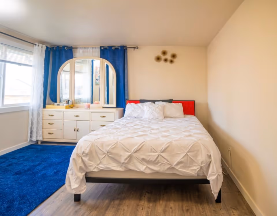 A bedroom with a bed covered in a white textured comforter and several pillows. Behind the bed is a wall with a small decorative piece. To the left of the bed is a white dresser with multiple drawers and a large mirror with three panels. Blue and white curtains frame a window next to the dresser. The floor is a combination of wood and a blue carpet.