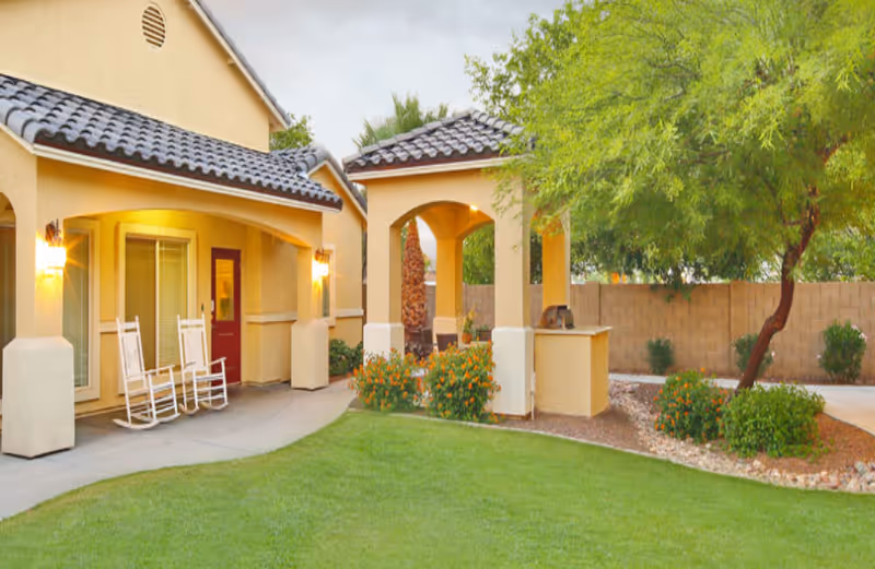 Outdoor view of a residential assisted living facility with a covered patio area featuring two white rocking chairs, a small garden with flowers, a tree, and a barbecue grill under an archway. The building has beige walls and a tiled roof.