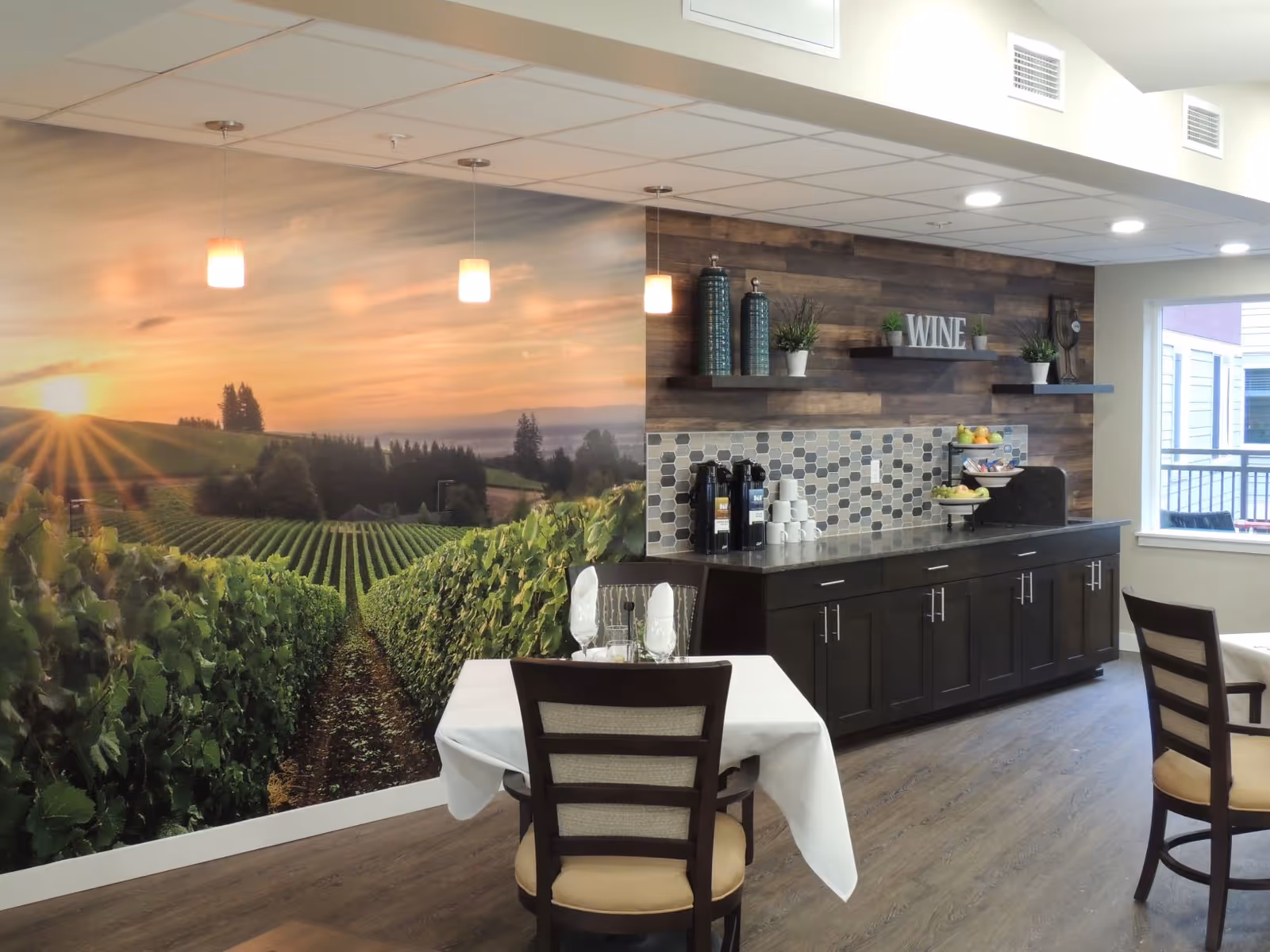 Dining area with tables and chairs, a buffet counter and coffee station beneath shelving and a 'WINE' sign, and a large vineyard mural on the wall.