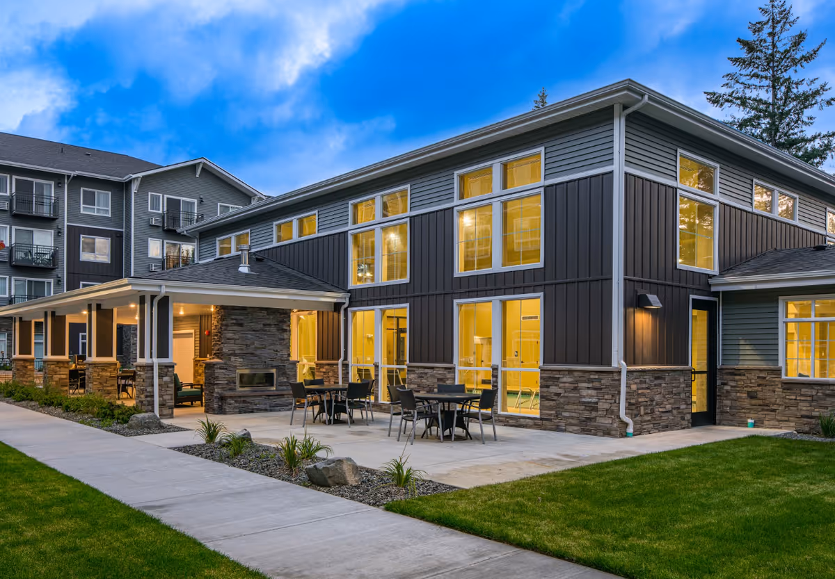 Exterior view of a modern senior living facility building at dusk with large windows illuminated from inside. The building features a covered patio area with outdoor seating and a fireplace, surrounded by a well-maintained lawn and sidewalk.