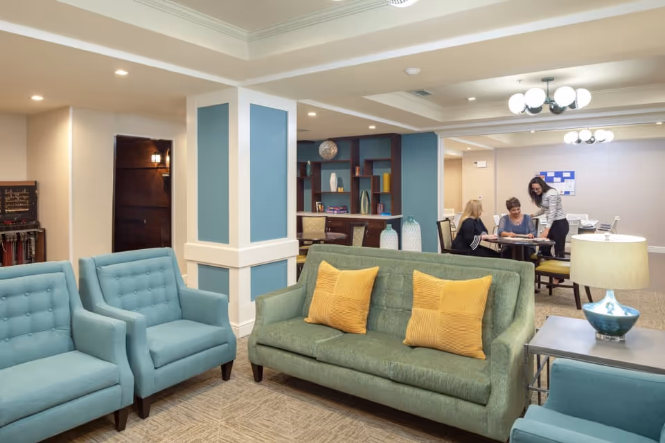 A cozy senior living facility common area with teal armchairs and a green sofa with yellow pillows. In the background, three women are seated at a table engaged in conversation or an activity. The room features soft lighting, a blue accent wall with shelves, and modern ceiling light fixtures.
