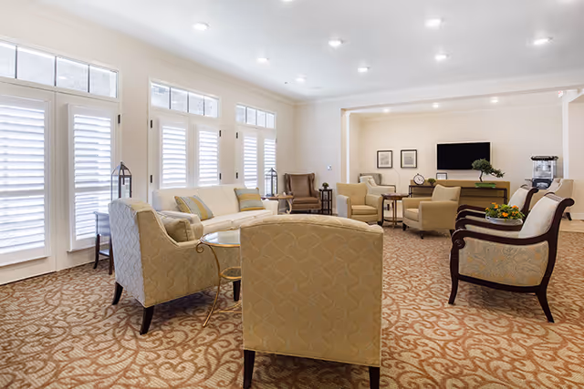A spacious and well-lit senior living common area with multiple upholstered armchairs and sofas arranged around small tables. The room features large windows with white plantation shutters, a patterned carpet, recessed ceiling lights, and a wall-mounted flat-screen TV. Decorative elements include framed pictures and a small plant on a side table.