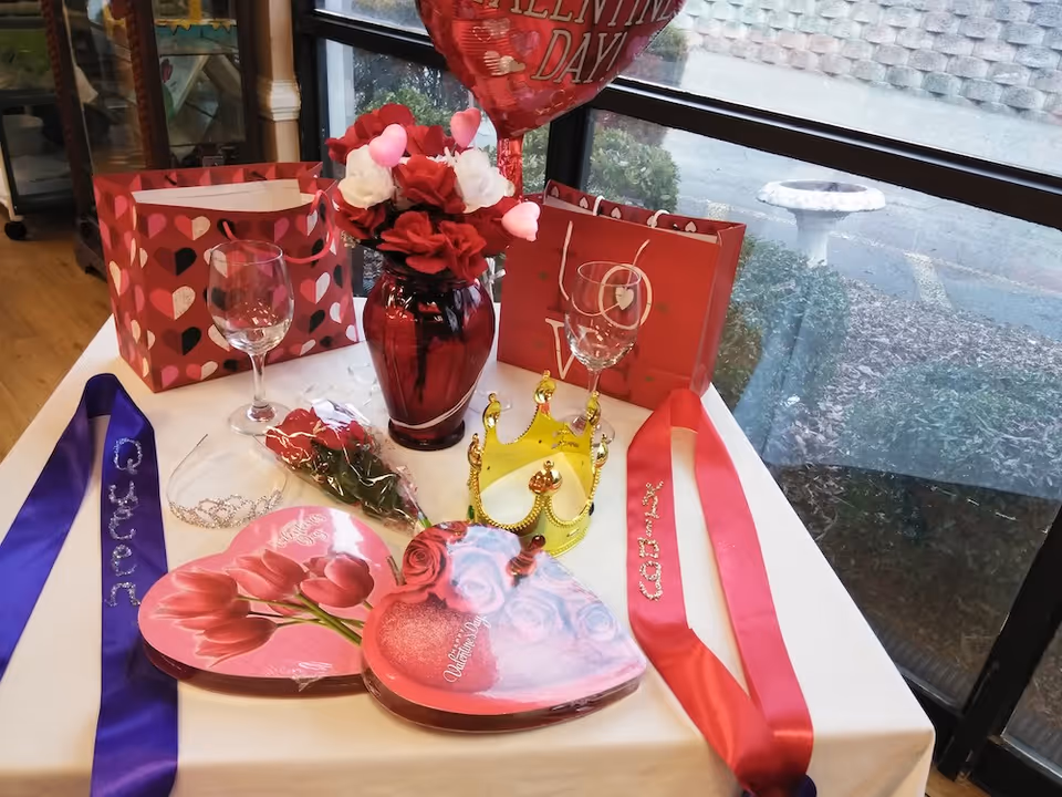 Valentine's Day display on a table with heart-shaped boxes, a red vase of flowers, wine glasses, gift bags, a crown, sashes, and a balloon by a window.