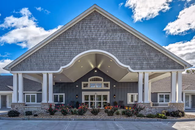 Front exterior view of Villas of Holly Brook Assisted Living & Memory Care building with a large covered entrance, white columns, stone accents, and landscaped flower beds under a partly cloudy blue sky.