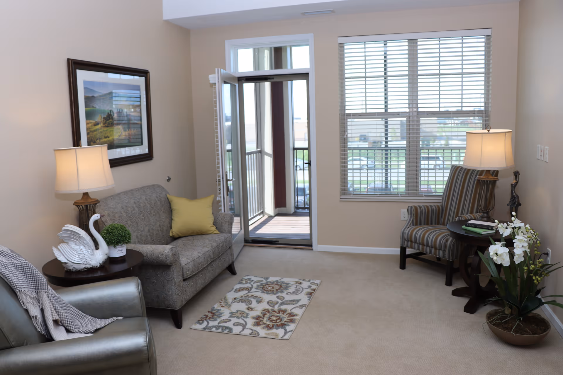 A cozy living room with beige walls and carpet. The room features a patterned loveseat with a yellow pillow, a metallic armchair with a gray throw, and a striped armchair. There are two side tables with lamps, a white swan figurine, a small green plant, and a decorative statue. A framed landscape picture hangs on the wall. A glass door opens to a small balcony, and a large window with blinds lets in natural light.