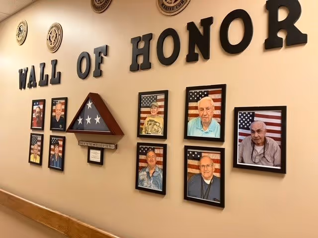 A Wall of Honor display in a hallway featuring framed photographs of veterans in front of American flags, a folded American flag in a triangular wooden case, and military emblems mounted above the display.