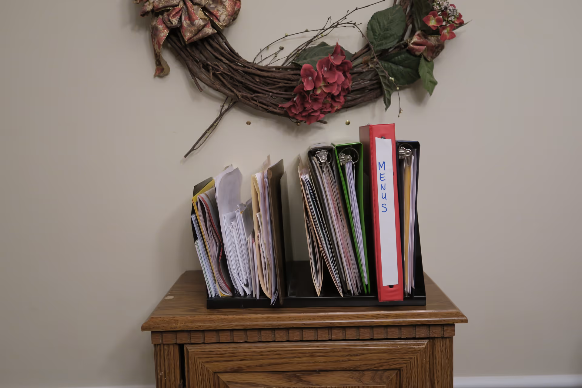 A wooden cabinet topped with an organizer full of folders and a red binder labeled "MENUS" beneath a decorative floral wreath on a plain wall.