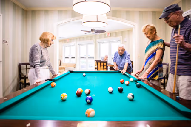 Four elderly people playing pool together in a well-lit room with large windows and comfortable seating in the background.