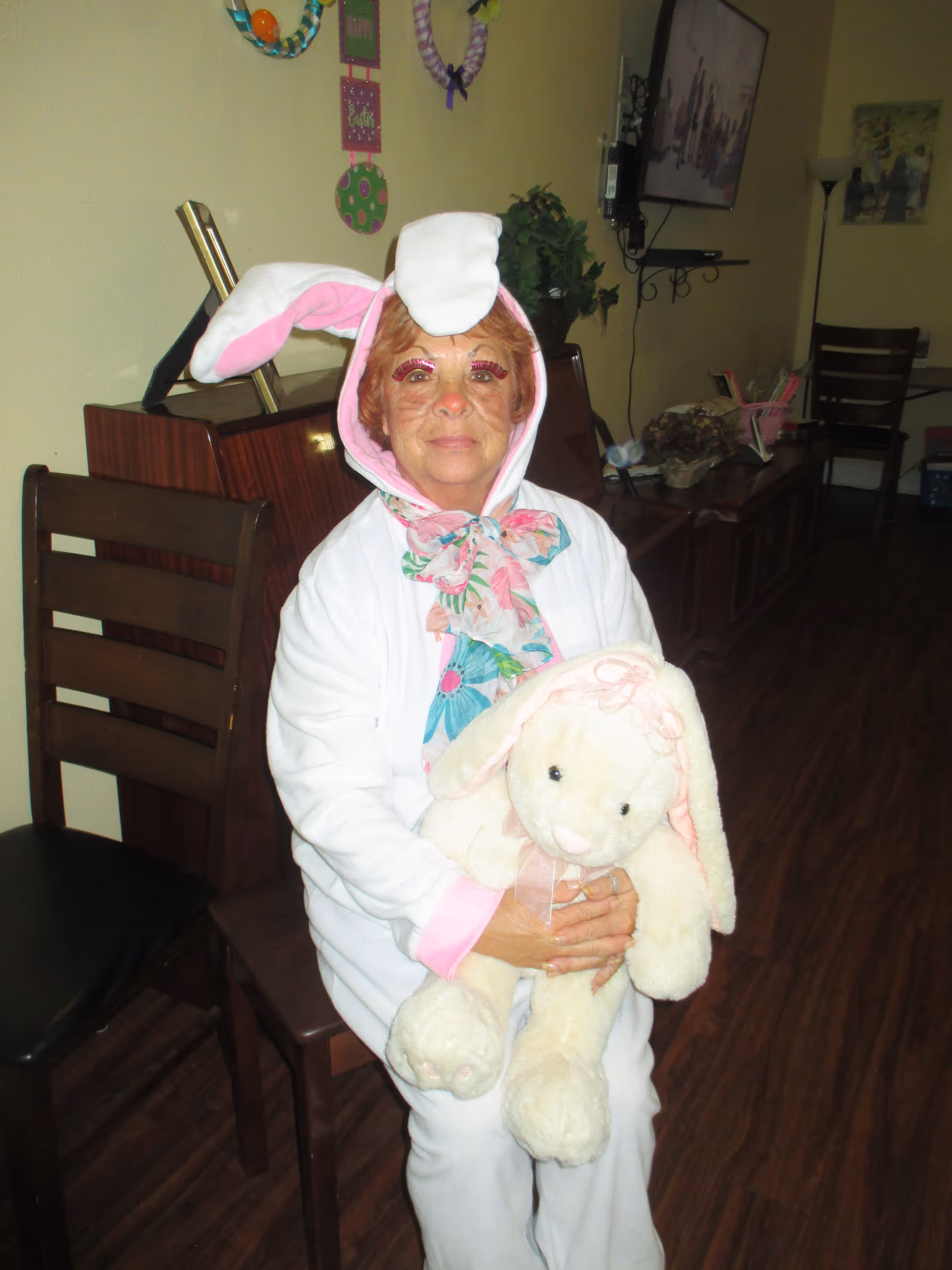 An elderly woman dressed in a white bunny costume with pink accents and a floral scarf is sitting on a wooden chair indoors, holding a large white stuffed bunny. The room has wooden flooring, a piano, chairs, and a TV mounted on the wall in the background.