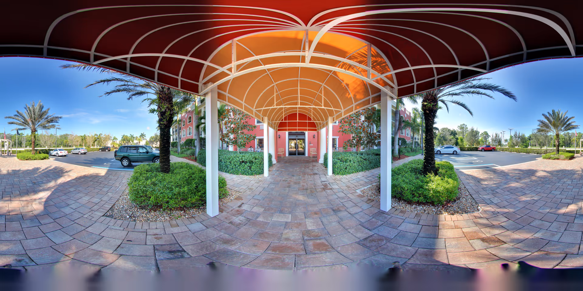 Covered walkway with a red and orange canopy leading to the entrance of a building. The pathway is paved with stone tiles and flanked by green shrubs and palm trees. Cars are parked in the parking lot on both sides under a clear blue sky.