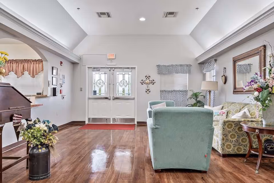 Bright reception lobby with mint-green armchairs, a patterned sofa, floral arrangements, and double glass entry doors.