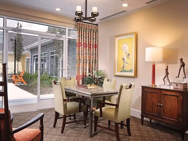 A cozy dining area with a square table and four upholstered chairs. The table has a chessboard and a plant centerpiece. There is a large window with patterned curtains letting in natural light and showing an outdoor patio with a bench. A wooden sideboard with a lamp and decorative sculptures is against the wall, which also features a framed artwork.