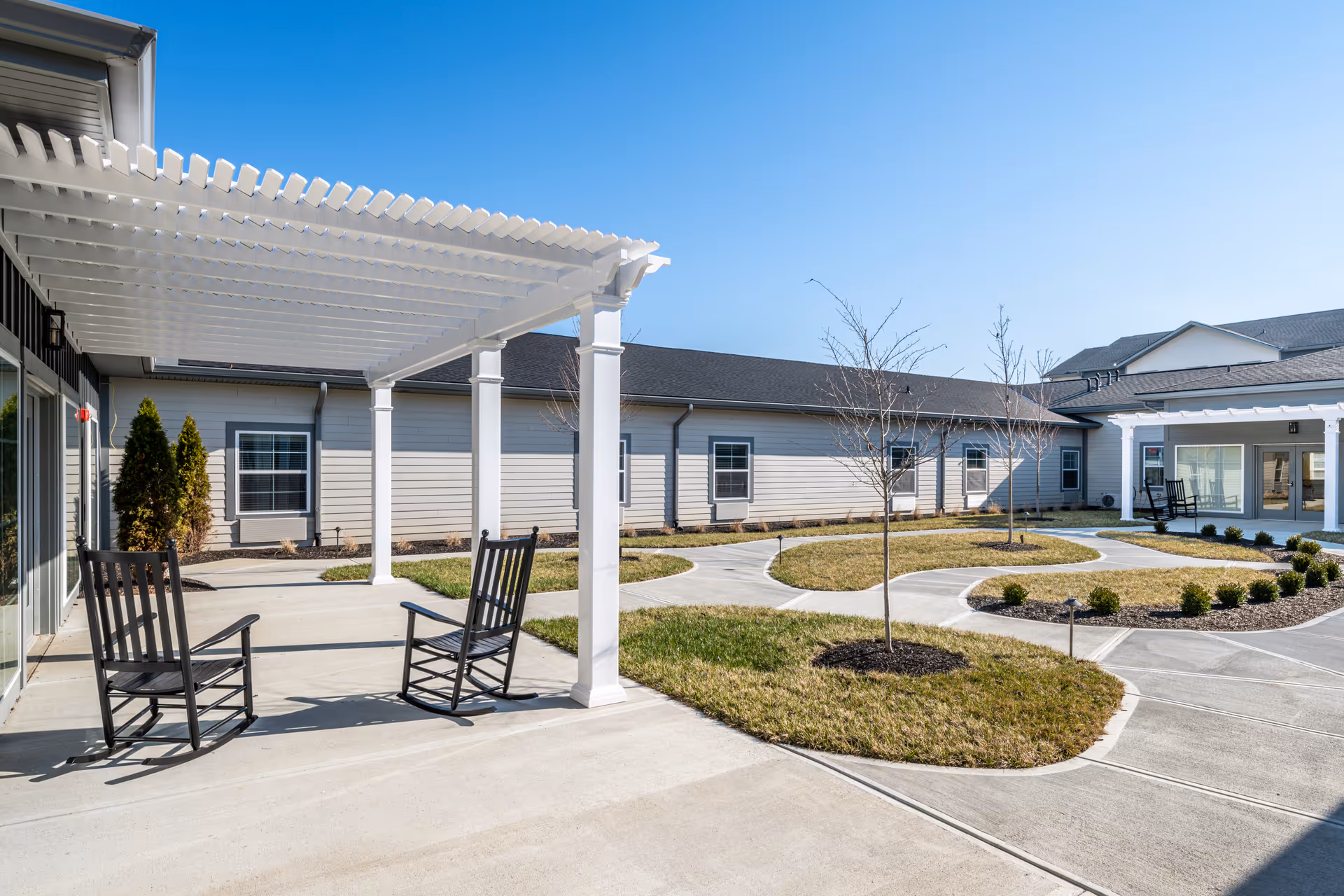 Sunlit courtyard with a white pergola, two black rocking chairs, paved winding paths, small landscaped islands, and a surrounding single-story building.