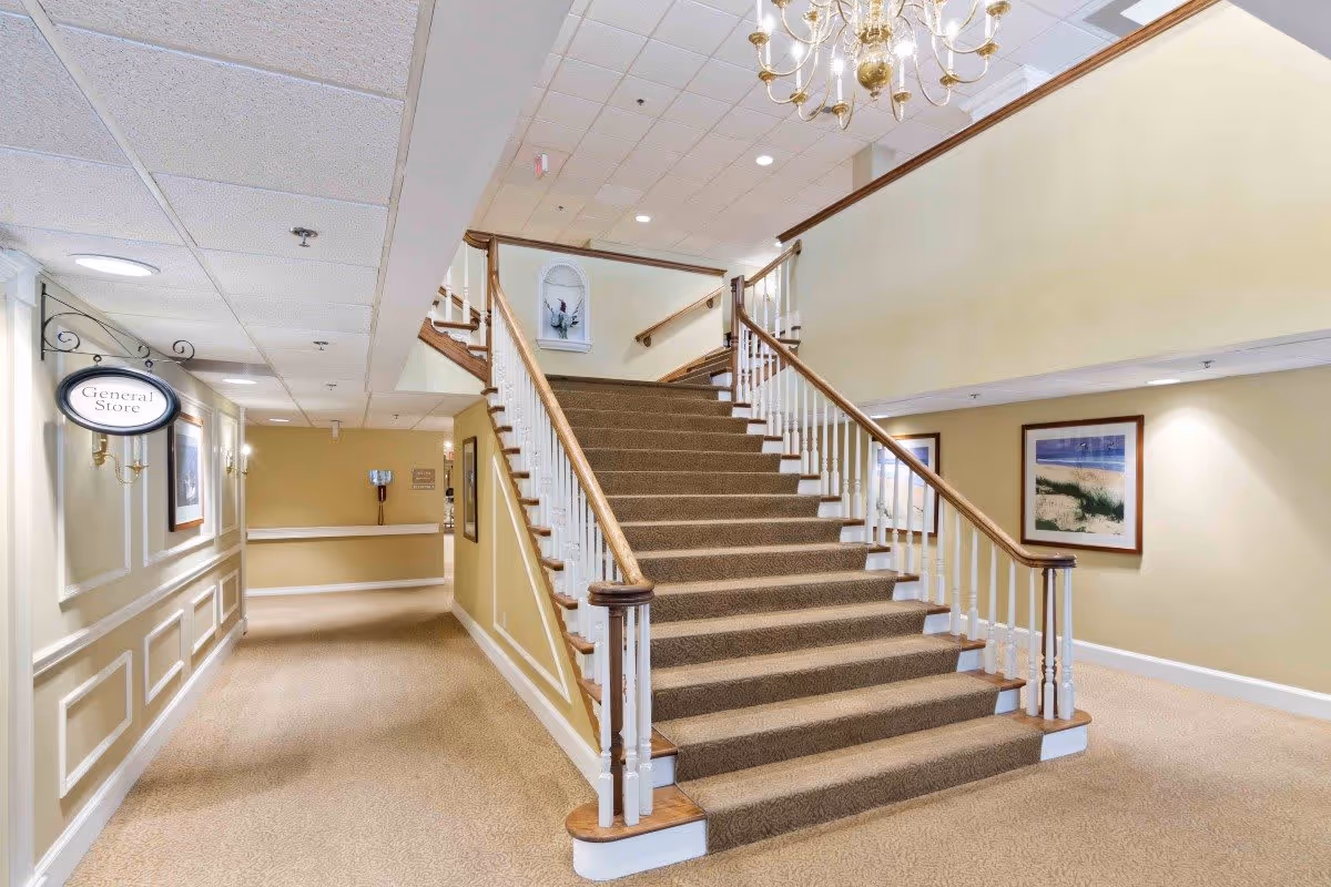 Interior view of a senior living facility hallway with a carpeted staircase leading to an upper floor. The walls are painted beige with white trim, and there are framed pictures hanging on the walls. A chandelier hangs from the ceiling above the staircase. A sign on the left side indicates the direction to the General Store.