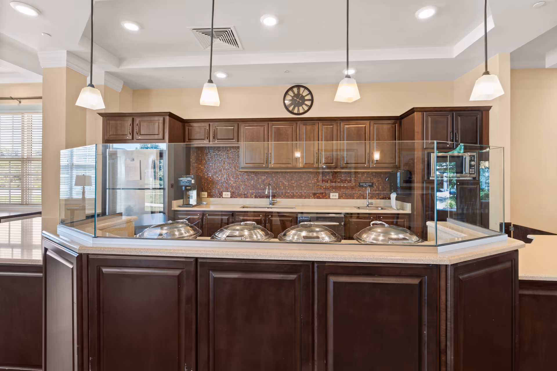 A clean and modern kitchen area with dark wood cabinets, a countertop with four covered serving dishes behind a glass sneeze guard, pendant lights hanging from the ceiling, a clock on the wall, and kitchen appliances including a refrigerator, coffee machine, and microwave.