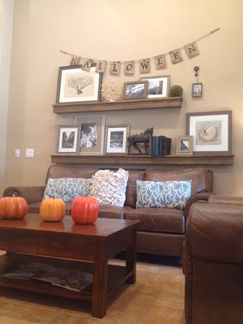 A cozy living room with a brown leather sofa adorned with decorative pillows. Above the sofa are two wooden shelves holding framed pictures, books, and decorative items. A banner spelling 'HALLOWEEN' hangs on the wall above the shelves. A wooden coffee table in front of the sofa has three small pumpkins placed on it.