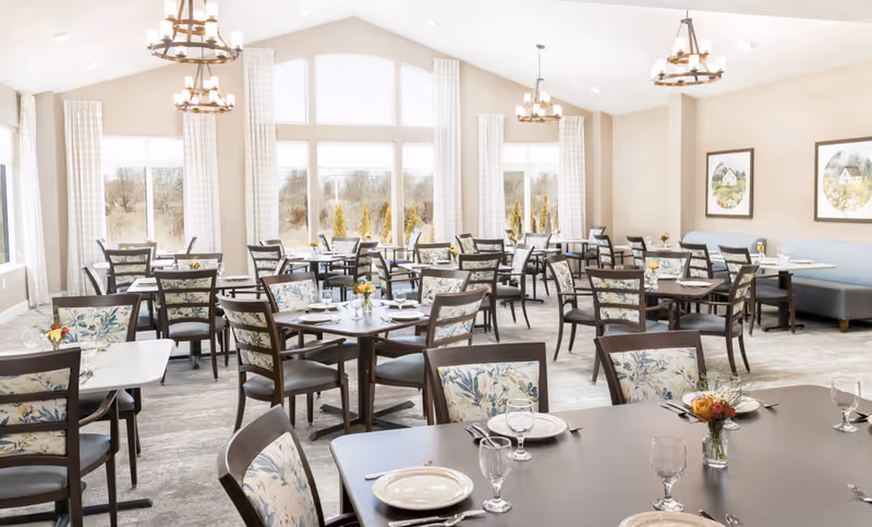 A bright and spacious dining room with multiple tables and chairs arranged neatly. The chairs have floral patterned cushions, and the tables are set with plates, glasses, and silverware. Large windows with white curtains allow natural light to fill the room. There are chandeliers hanging from the ceiling and framed artwork on the walls.