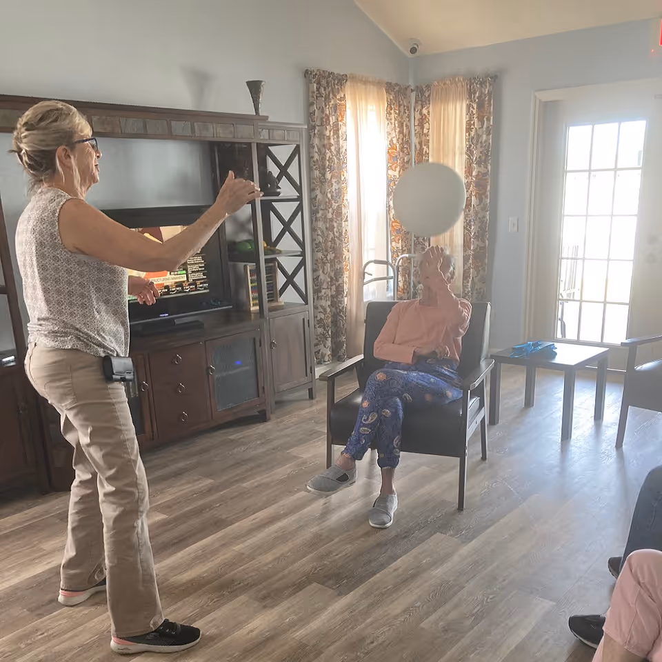 An elderly woman sitting in a chair indoors, playing with a white balloon, while another woman stands nearby engaging with her. The room has wooden flooring, a large entertainment center with a TV, floral curtains, and a glass door letting in natural light.
