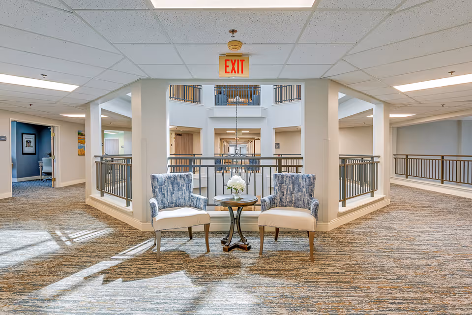 A bright and spacious hallway area in a senior living facility with two blue and white patterned armchairs and a small round table with a flower arrangement between them. The hallway has carpeted floors, white walls, and a railing overlooking a lower level. An exit sign hangs from the ceiling above the seating area.
