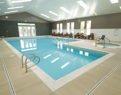 Indoor swimming pool area with clear blue water, surrounded by beige tiled flooring. Several chairs are lined up against the far wall with large windows letting in natural light. There is also a hot tub to the right side of the pool. The ceiling is high with multiple skylights and recessed lighting.