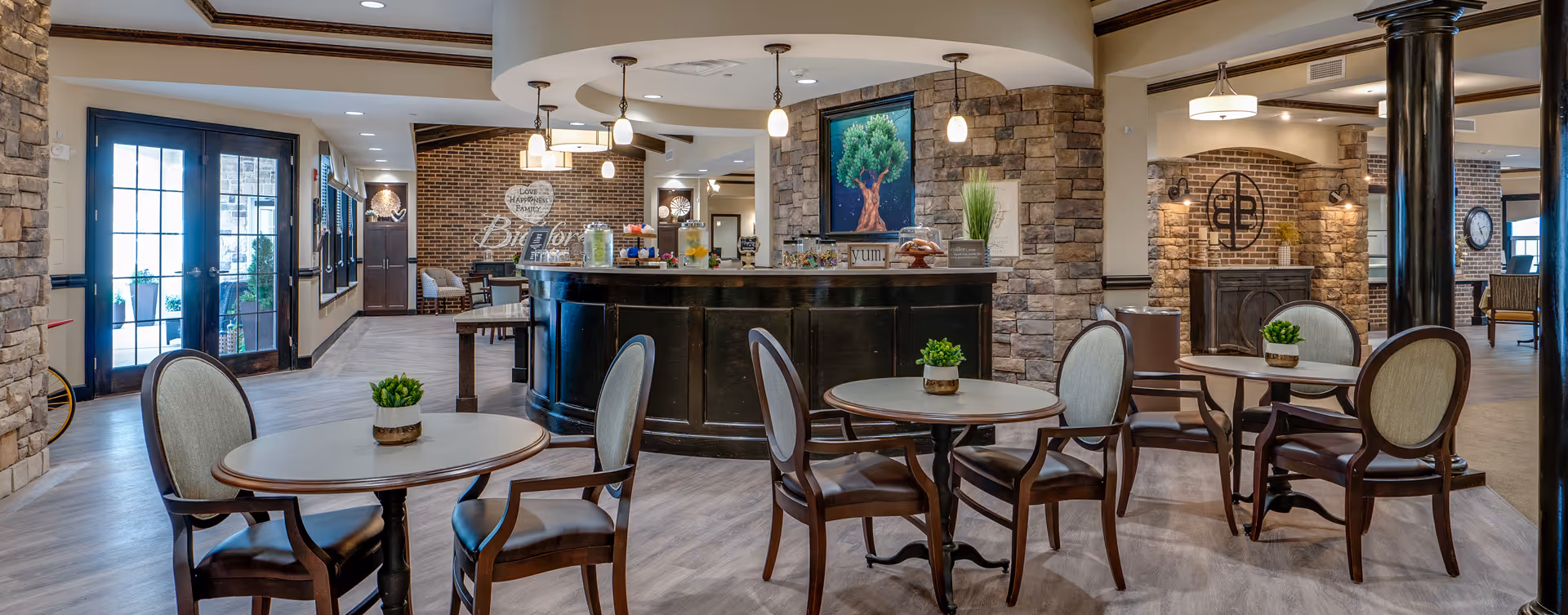 Bright communal lounge with round tables and chairs facing a central reception/coffee bar and stone-accented walls.