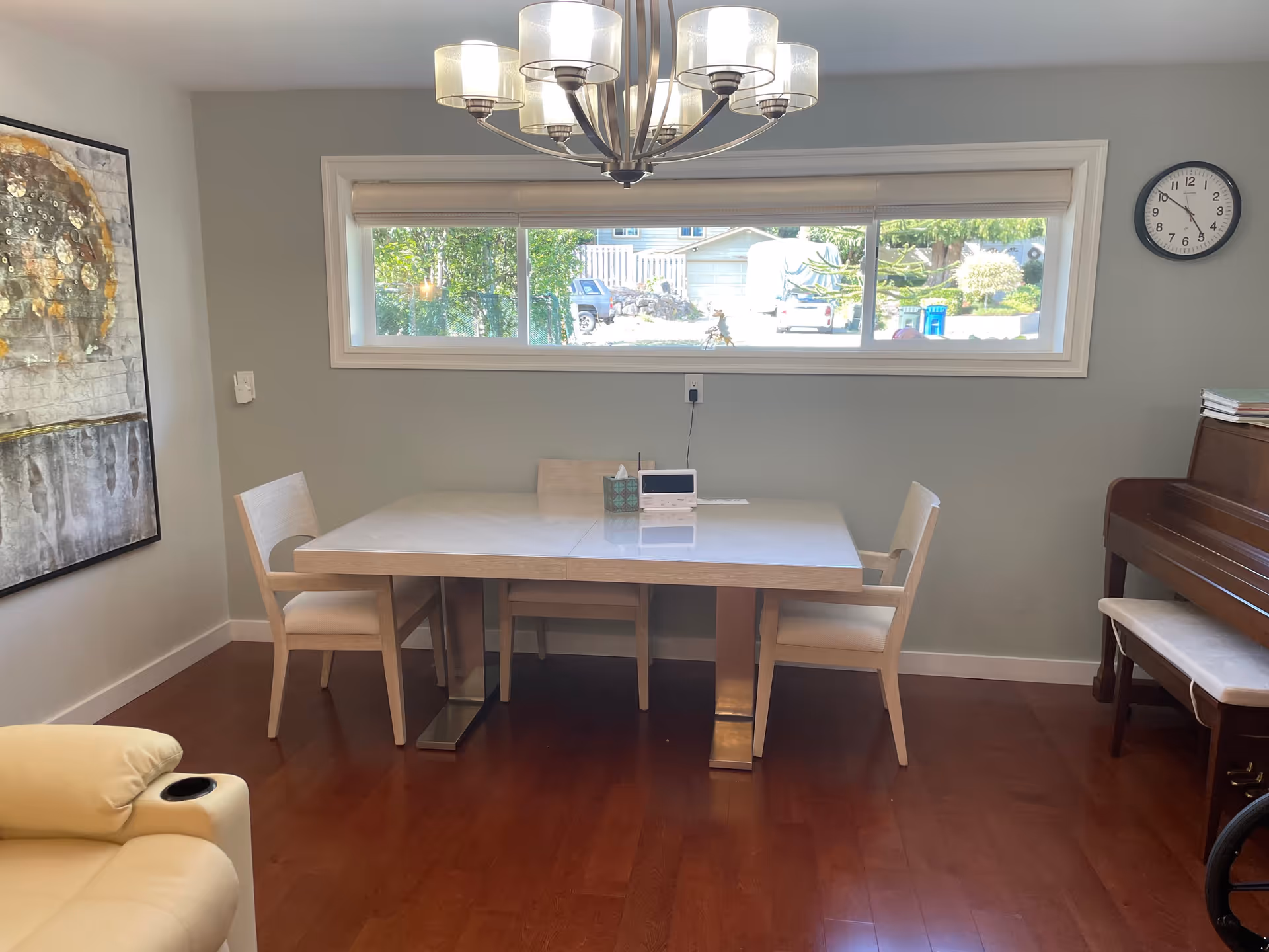 A dining area with a rectangular table and four chairs. Above the table hangs a modern chandelier with multiple lights. A large horizontal window behind the table shows a view of the outdoors with trees and parked vehicles. To the right, there is a piano with a bench, and a wall clock is mounted above it. On the left wall, there is a large abstract painting. The floor is wooden, and a beige armchair is partially visible in the foreground.