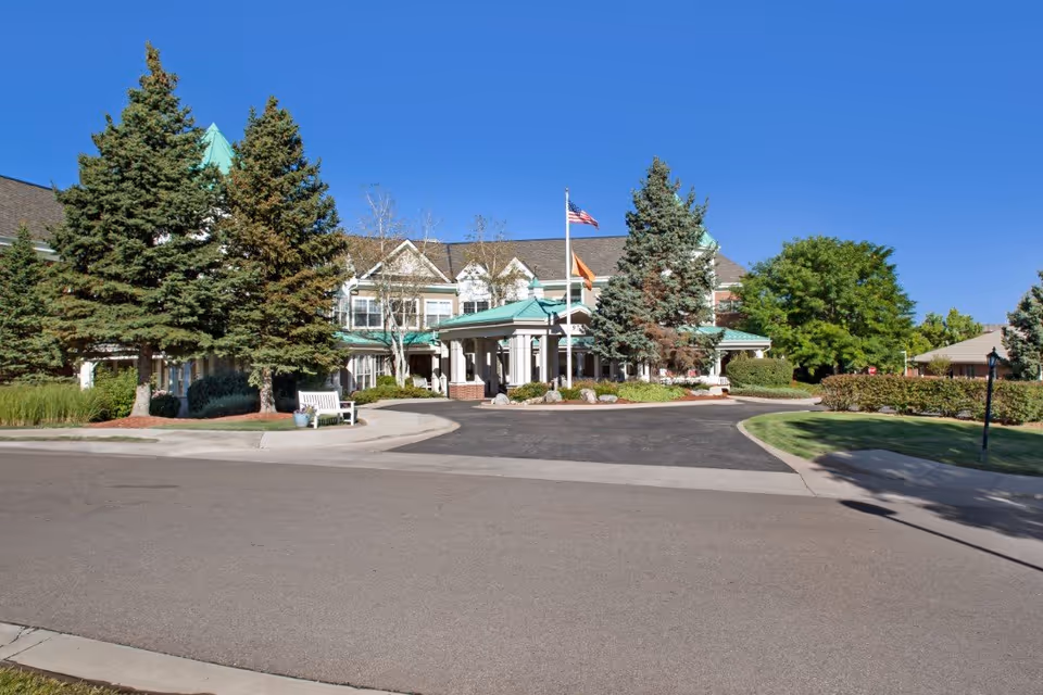 Front exterior view of a senior living facility named Sunrise at Orchard with a circular driveway, several large evergreen trees, a flagpole with an American flag, and a clear blue sky.