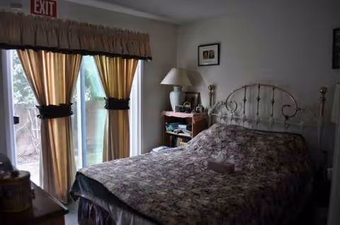 A furnished bedroom with floral bedspread, metal headboard, nightstand, lamp, and yellow-curtained sliding doors.