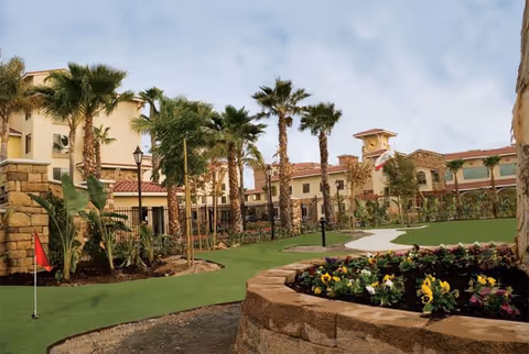 Outdoor landscaped area at Paradise Village featuring a putting green with a red flag, palm trees, a flower bed with colorful flowers, and a multi-story building with a tower in the background under a cloudy sky.
