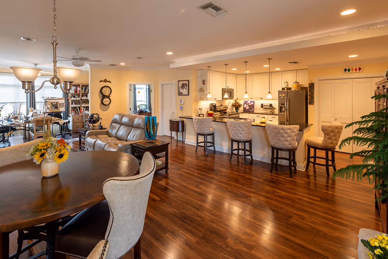 Spacious open-concept interior of a senior living facility showing a dining area with a round wooden table and upholstered chairs, a living area with a leather sofa, and a kitchen with a breakfast bar featuring four cushioned bar stools. The room has warm wooden flooring, soft yellow walls, pendant lighting over the kitchen counter, and various decorative items including flowers and framed pictures.
