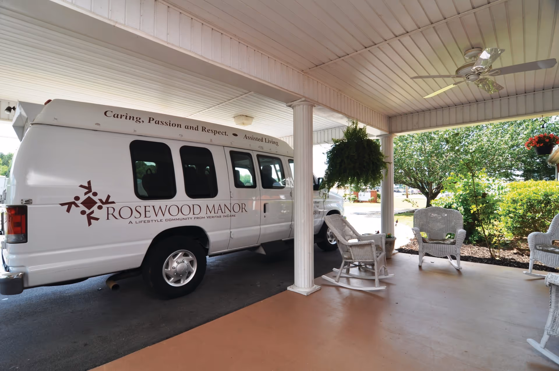 A white van with the logo and name Rosewood Manor parked under a covered porch area with white columns. The porch has white wicker rocking chairs, a hanging fern plant, and a ceiling fan. Green bushes and trees are visible in the background.