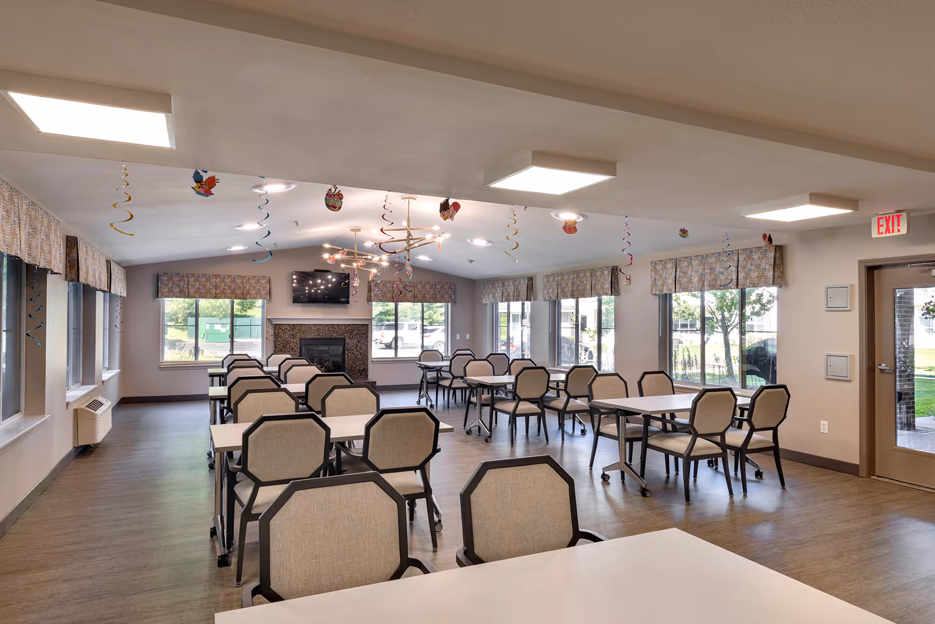A spacious dining room with multiple tables and chairs arranged neatly. The room has large windows with patterned valances allowing natural light to enter. There is a fireplace with a mounted TV above it on the far wall. Colorful spiral decorations hang from the ceiling, and there is an exit door on the right side leading outside.