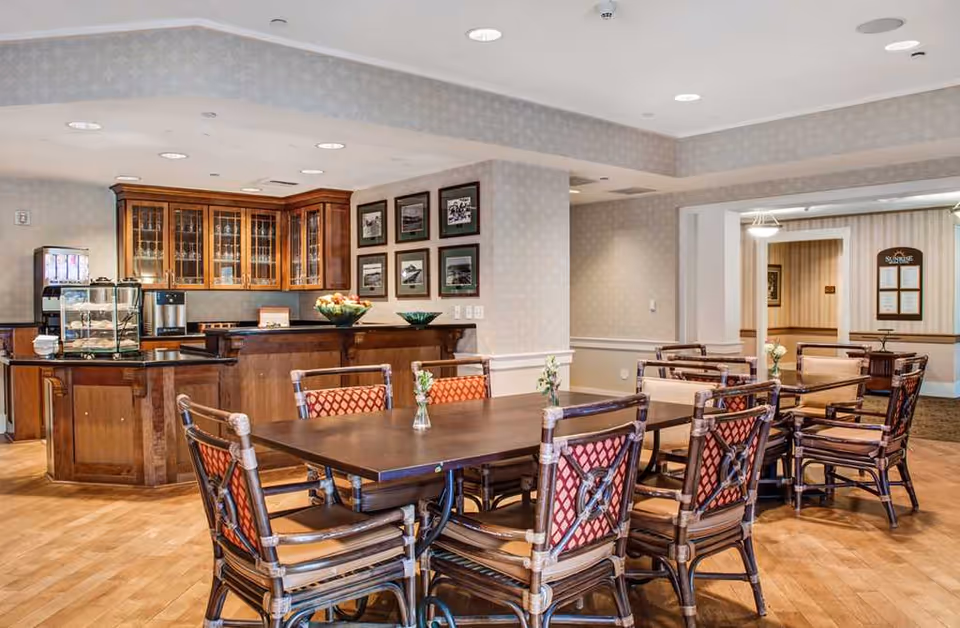 A dining area in a senior living facility featuring wooden tables and chairs with red patterned cushions. In the background, there is a wooden cabinet with glass doors and a counter with a fruit bowl and a coffee machine. The walls are decorated with framed pictures and the floor is wooden.