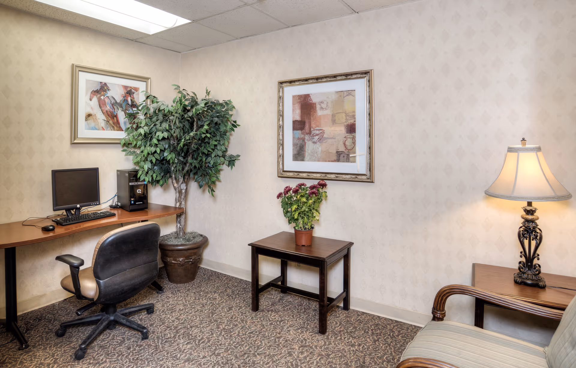 Interior sitting area with a computer workstation, potted plants, framed art, side table, lamp, and an upholstered chair.