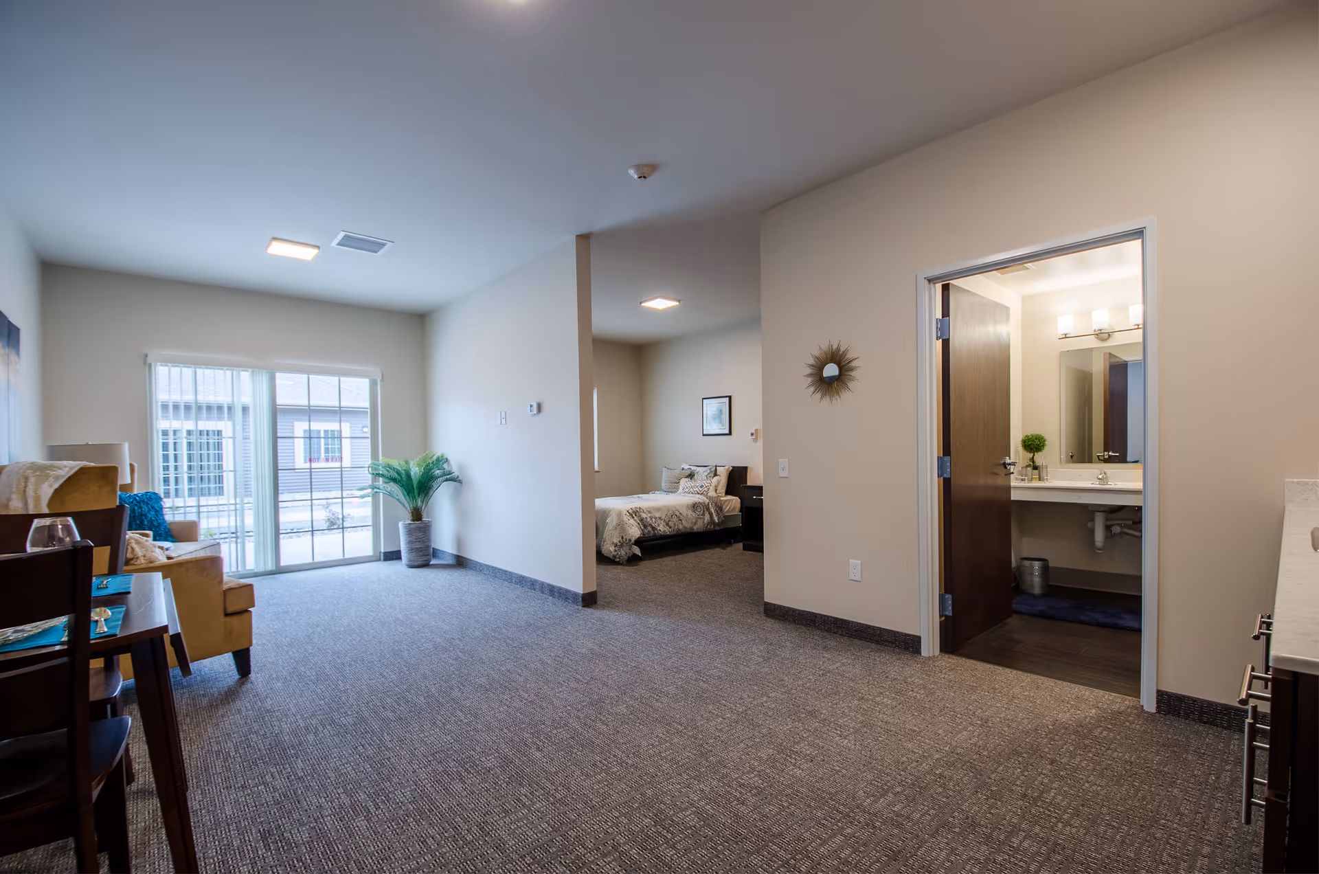 Interior view of a senior living facility apartment at Chesaning Comfort Care showing a living area with carpeted floor, a small dining table with chairs, a beige armchair, and a potted plant near a large window with vertical blinds. The image also shows a bedroom with a bed and nightstand, and a bathroom with a sink and mirror visible through an open door.