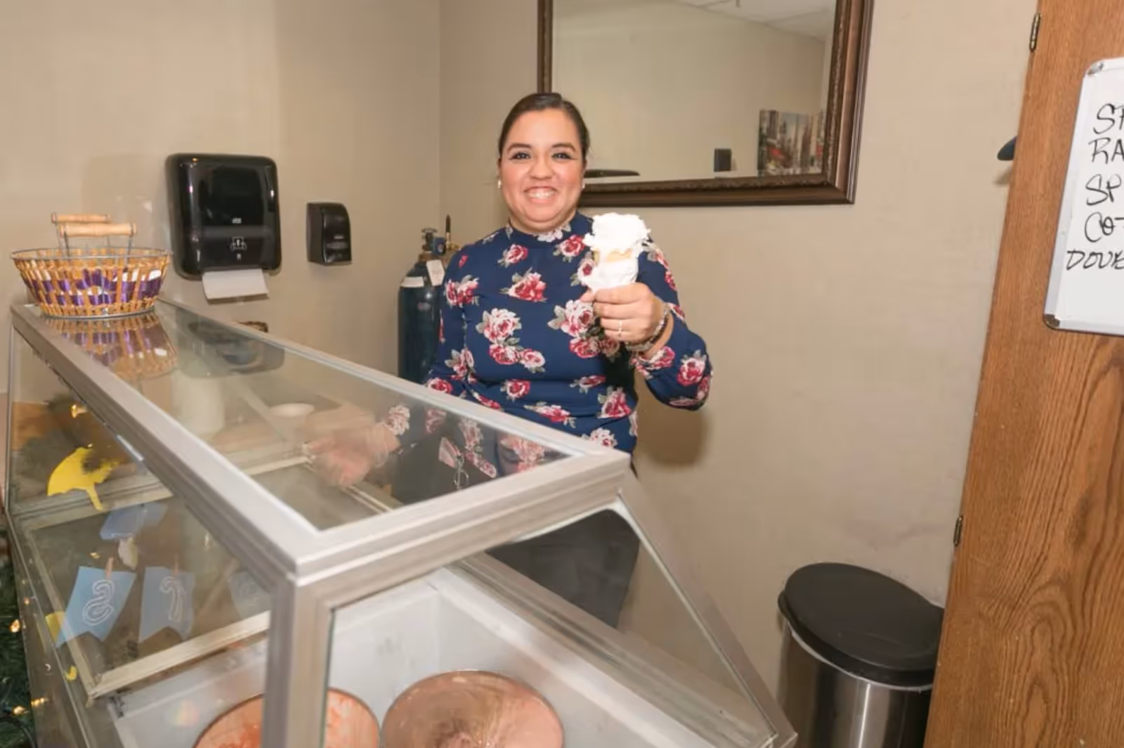 A smiling woman wearing a floral top stands behind a glass ice cream display case holding an ice cream cone with whipped cream. The setting appears to be indoors with a mirror on the wall behind her, a basket on the display case, and a trash can nearby.