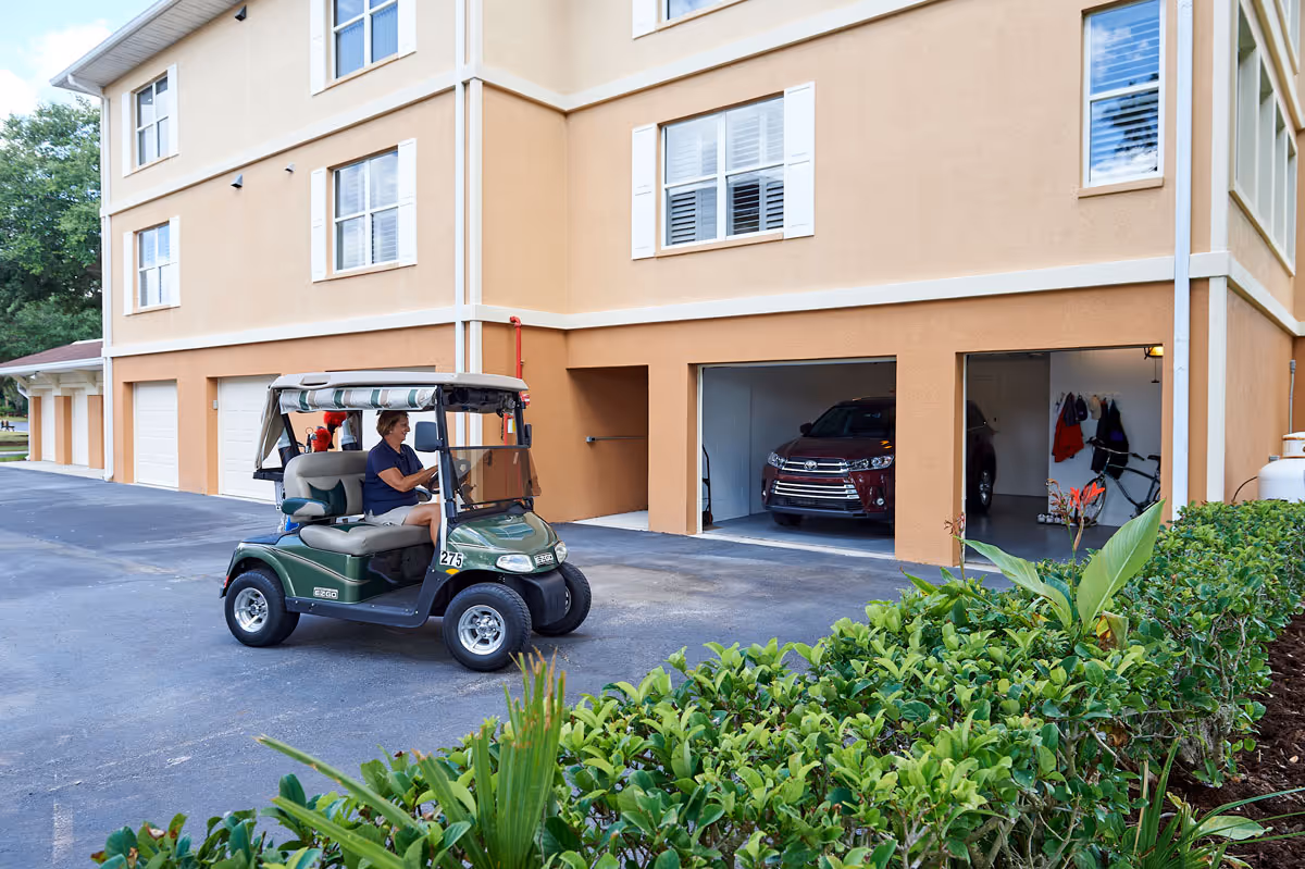 A person driving a green golf cart in front of a beige multi-story building with several garage doors, some open showing parked cars inside. There is greenery in the foreground and trees in the background.